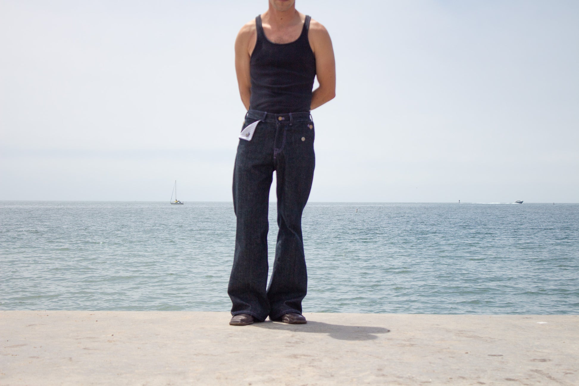 Person wearing black tank top and flared jeans standing by a body of water.